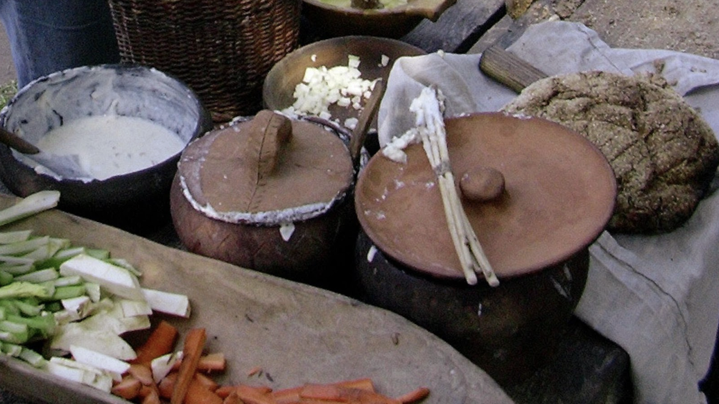 Image displays an assortment of traditional food preparation items. Visible are clay pots with lids, one containing a white dairy product, alongside baskets and wooden utensils. Chopped vegetables, including carrots and cucumbers, are laid out on a wooden tray